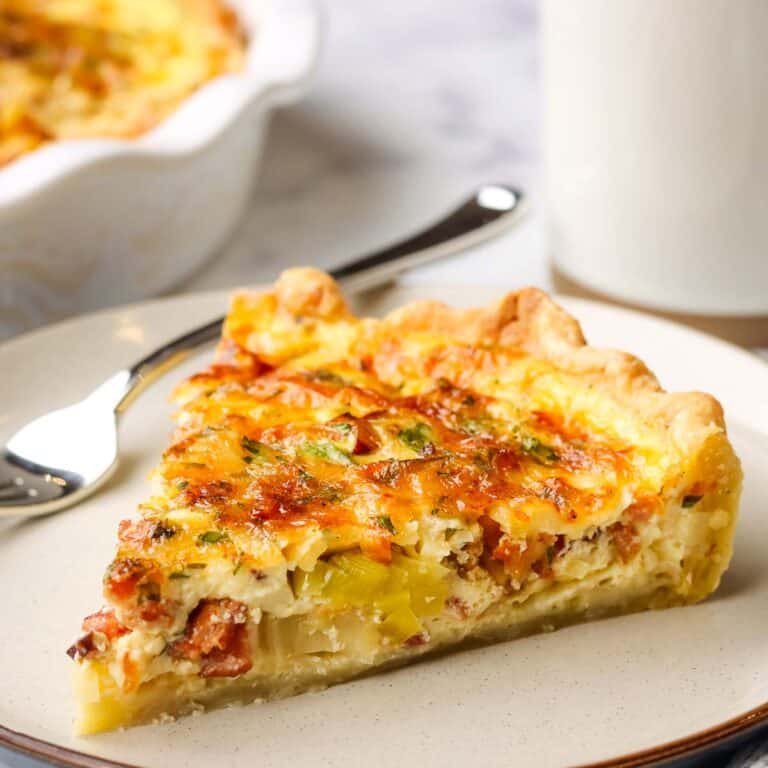 a slice of bacon and leek quiche with a dish of quiche and coffee mug in the background