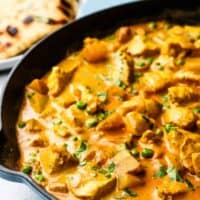 A cast iron pan full of chicken and potato curry garnished with cilantro, with a plate of naan bread in the background.