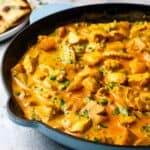 A cast iron pan full of chicken and potato curry garnished with cilantro, with a plate of naan bread in the background.