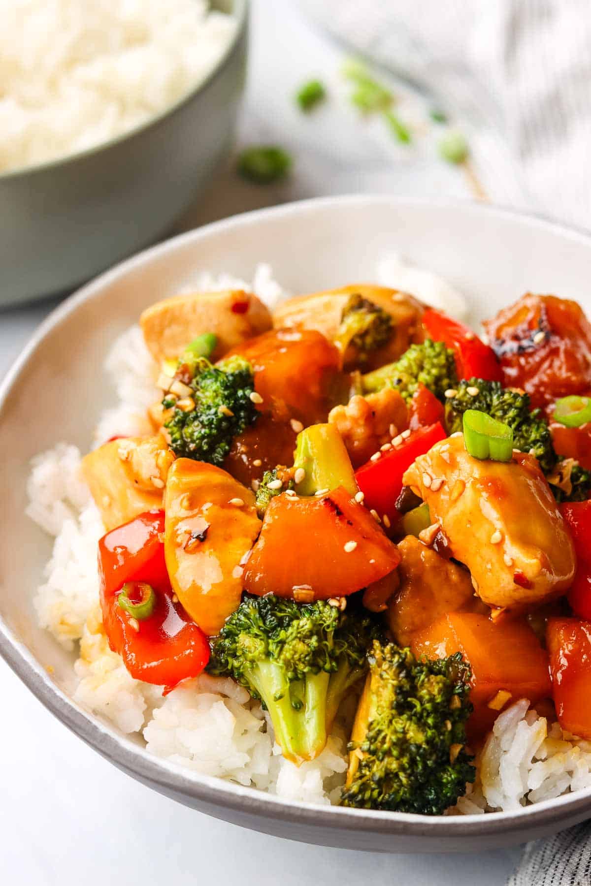 A bowl of chicken teriyaki with vegetables stir fry on a bed of rice with a bowl of rice in the background.