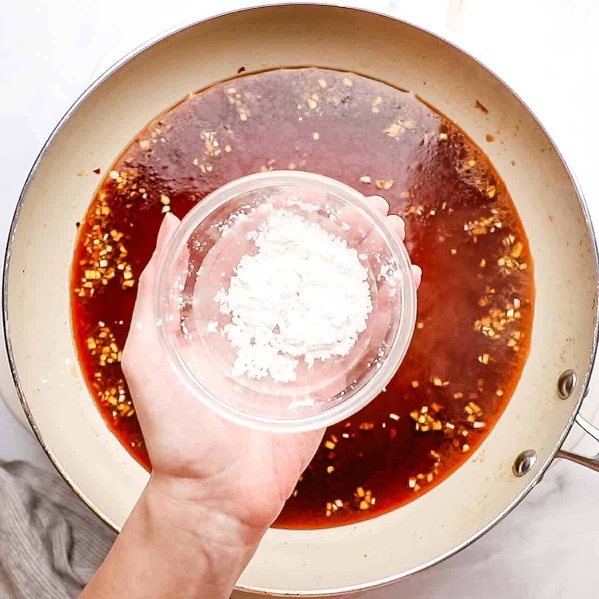 a hand holding a small bowl with cornstarch on top of a pan with teriyaki sauce in it