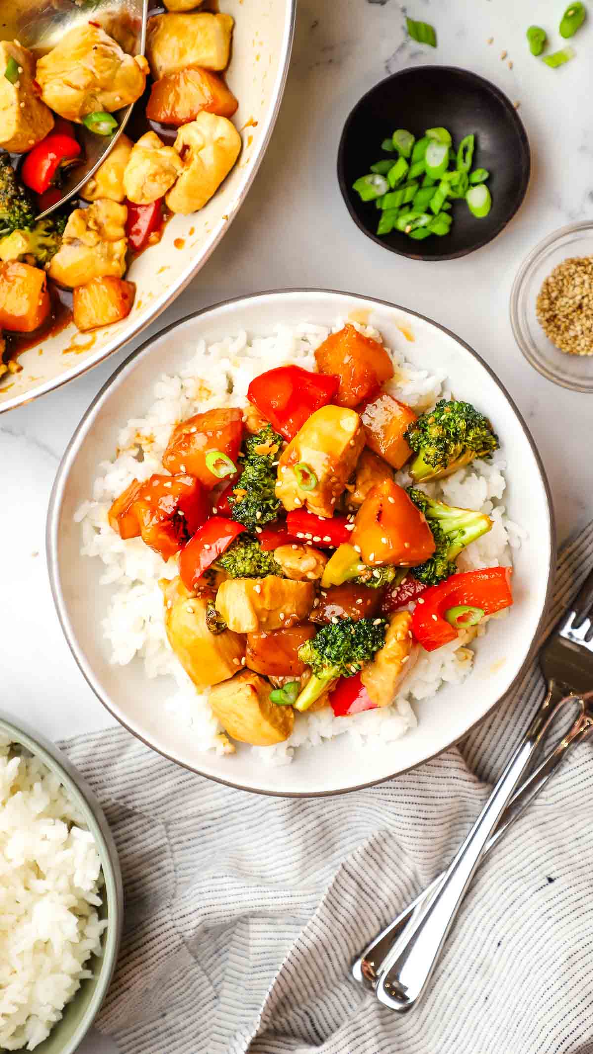 A bowl of chicken teriyaki with vegetables stir fry on a bed of rice with a pan of stir fry and a bowl of rice in the background.