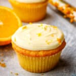 An orange cupcake topped with a swirl of orange cream cheese frosting and garnished with orange zest with oranges in the background.