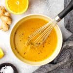 A bowl of ginger orange ginger dressing with a whisk in it and an orange half and ginger root in the background.