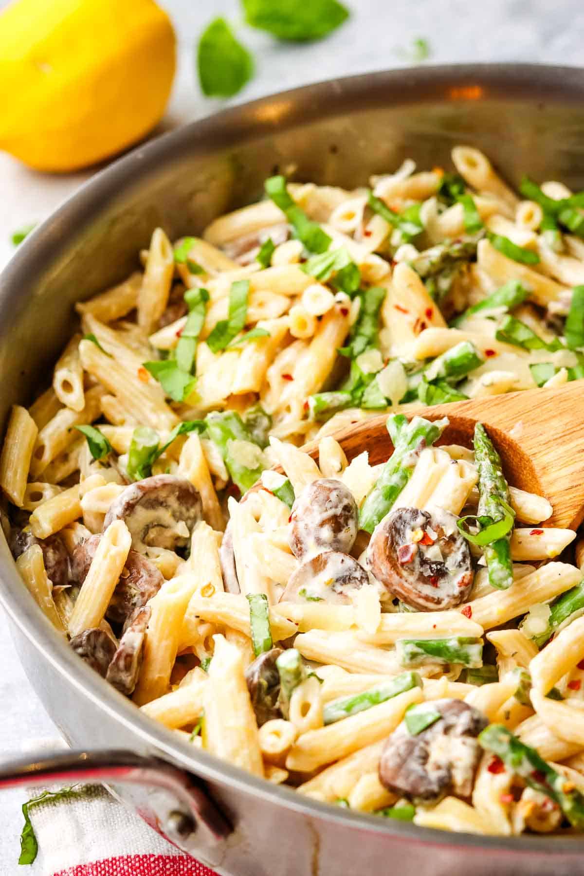 A pan of asparagus mushroom pasta with a wooden serving spoon in it.