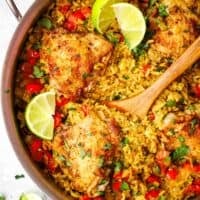 A pan of Cuban-style yellow rice with red peppers and chicken thighs garnished with limes.