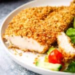 A piece of breaded chicken breast on a plate with a piece sliced off to show the inside.