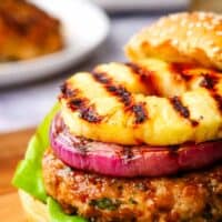 Closeup of a chicken burger topped with grilled red onion and grilled pineapple on a wooden serving board.