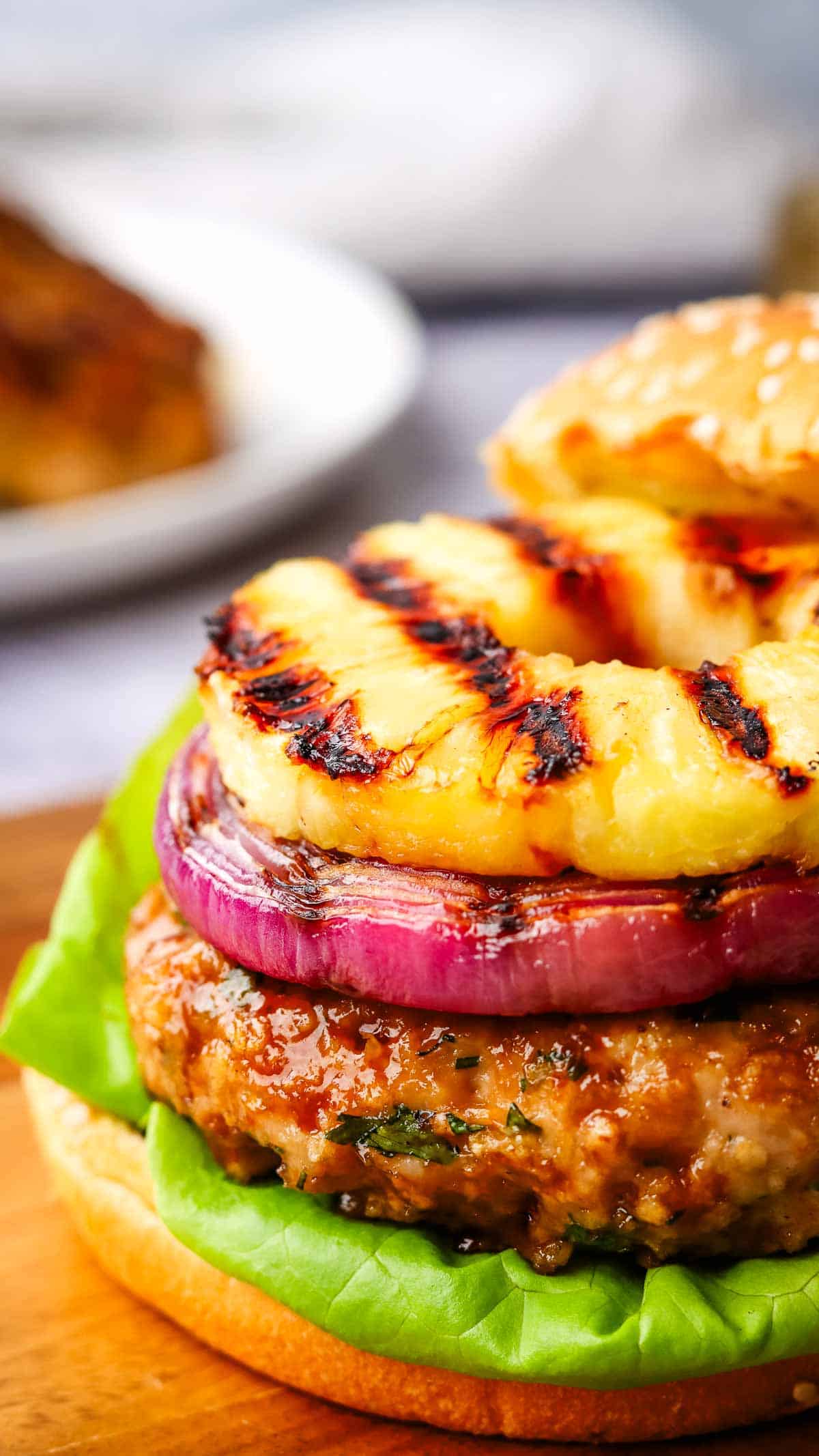 Closeup of a chicken burger topped with grilled red onion and grilled pineapple on a wooden serving board.