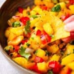 A bowl of peach mango salsa with a hand dipping a chip.
