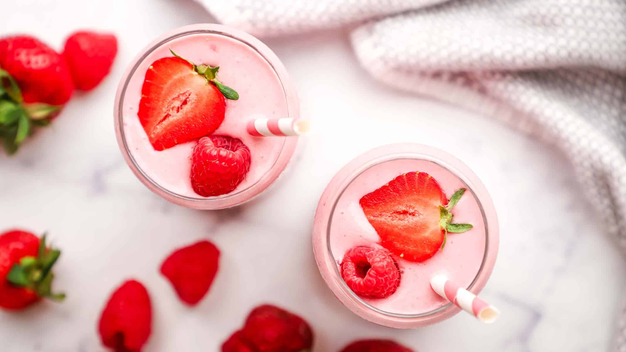 overhead view of two glasses of strawberry raspberry smoothie topped with fresh strawberries and with pink straws in the glasses.