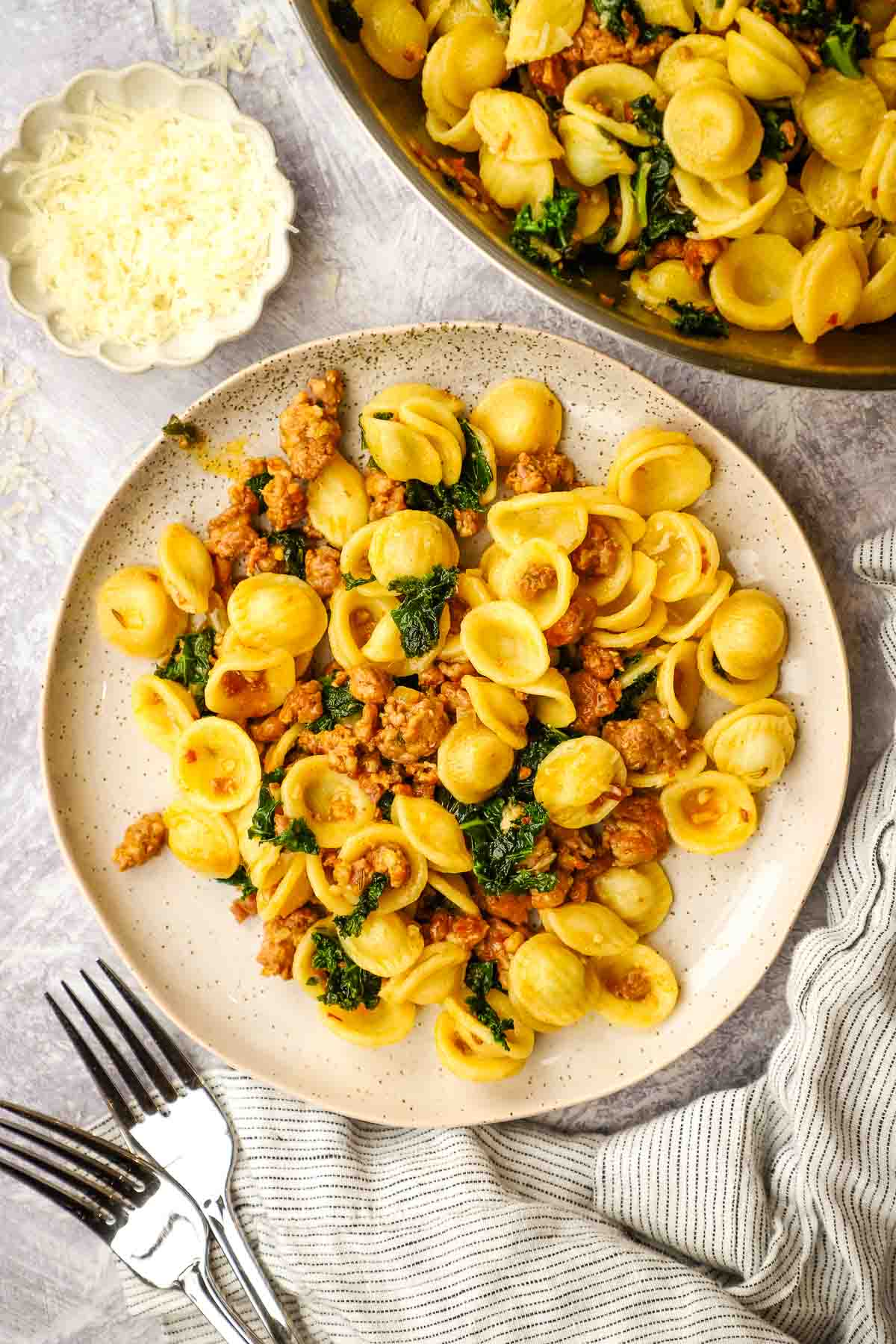 A plate of kale sausage pasta with a pan of pasta in the background.