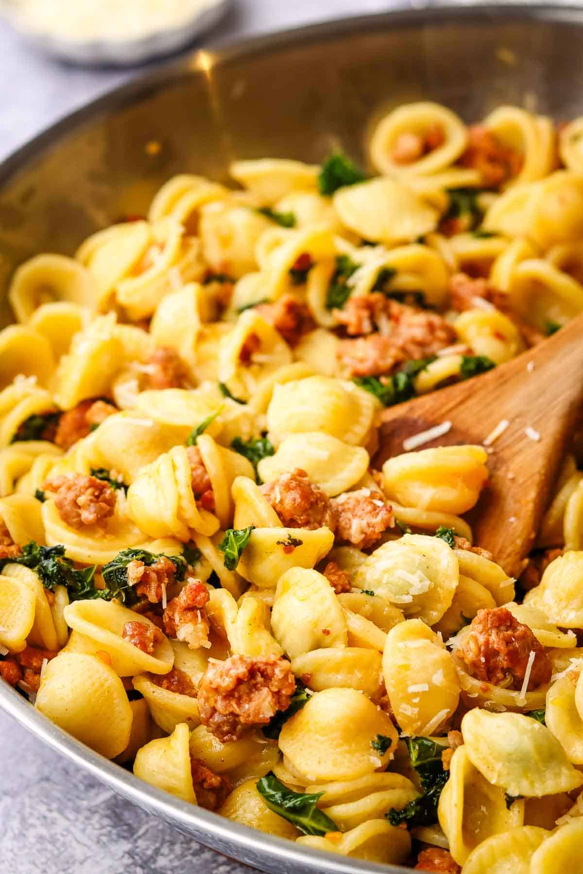 a pan of sausage kale pasta with a wooden serving spoon in it.