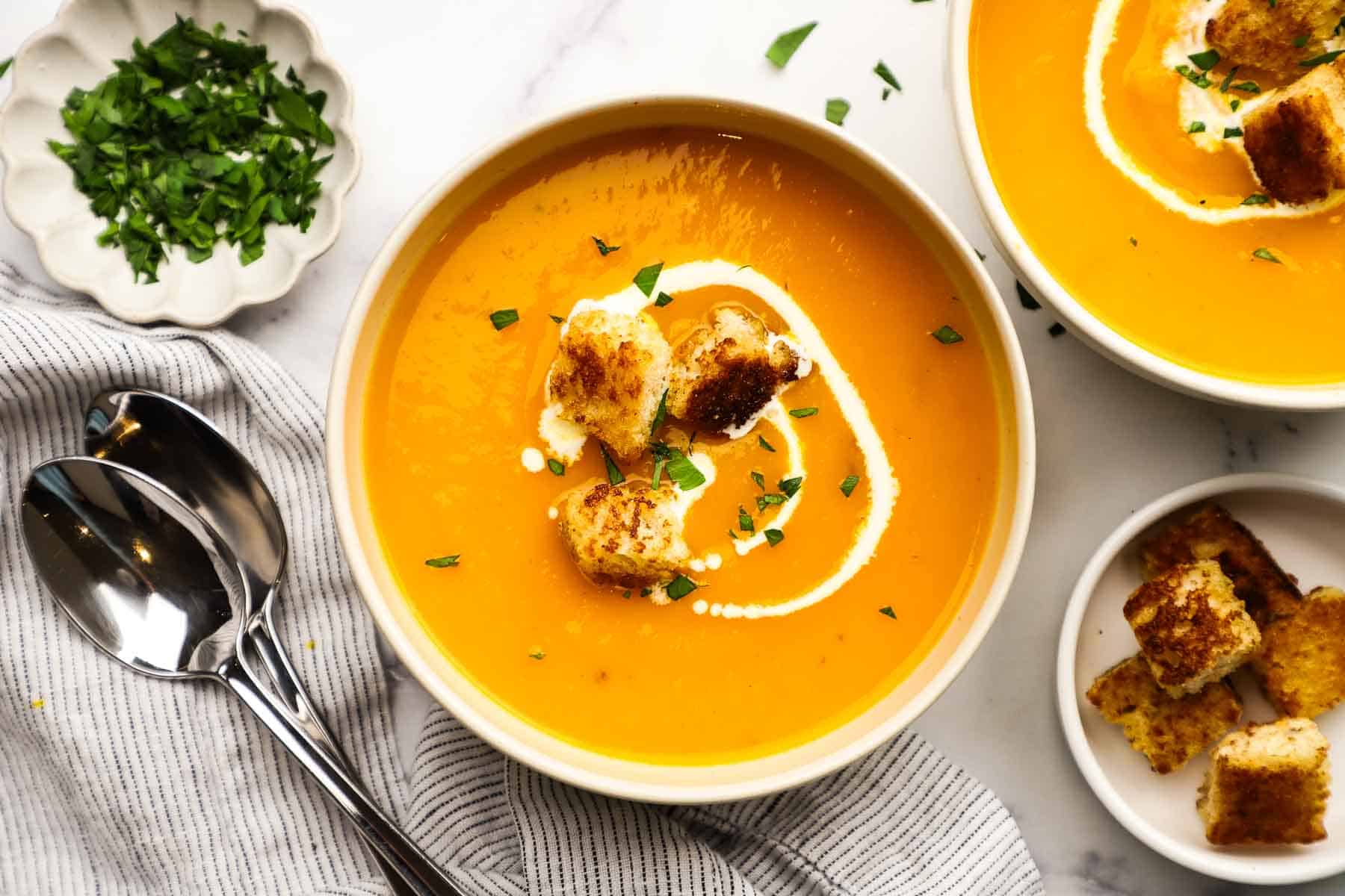 A bowl of butternut squash soup garnished with cream and croutons and with the toppings on the side.