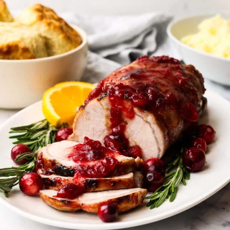 A cranberry pork loin roast on a serving platter with biscuits and mashed potatoes in the background.