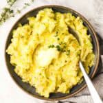 A bowl of mashed potatoes with butter on top and garnished with a thyme sprig with a spoon in it.