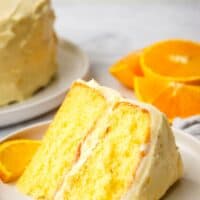A slice of a two layer orange cake on a plate with orange slices in the background.