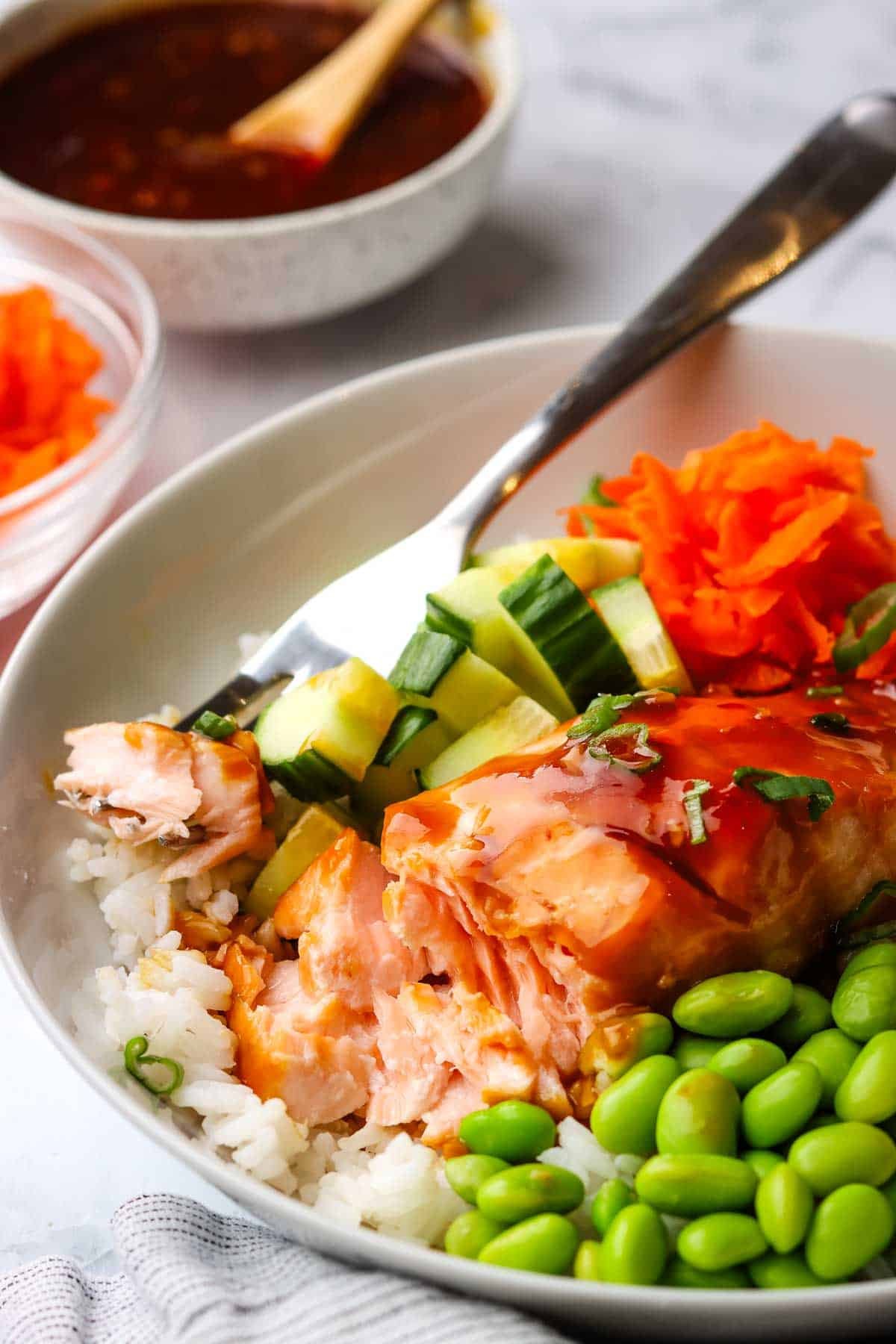 a fork taking a bite of the salmon from a bowl with rice, salmon, veggies, and sauce.