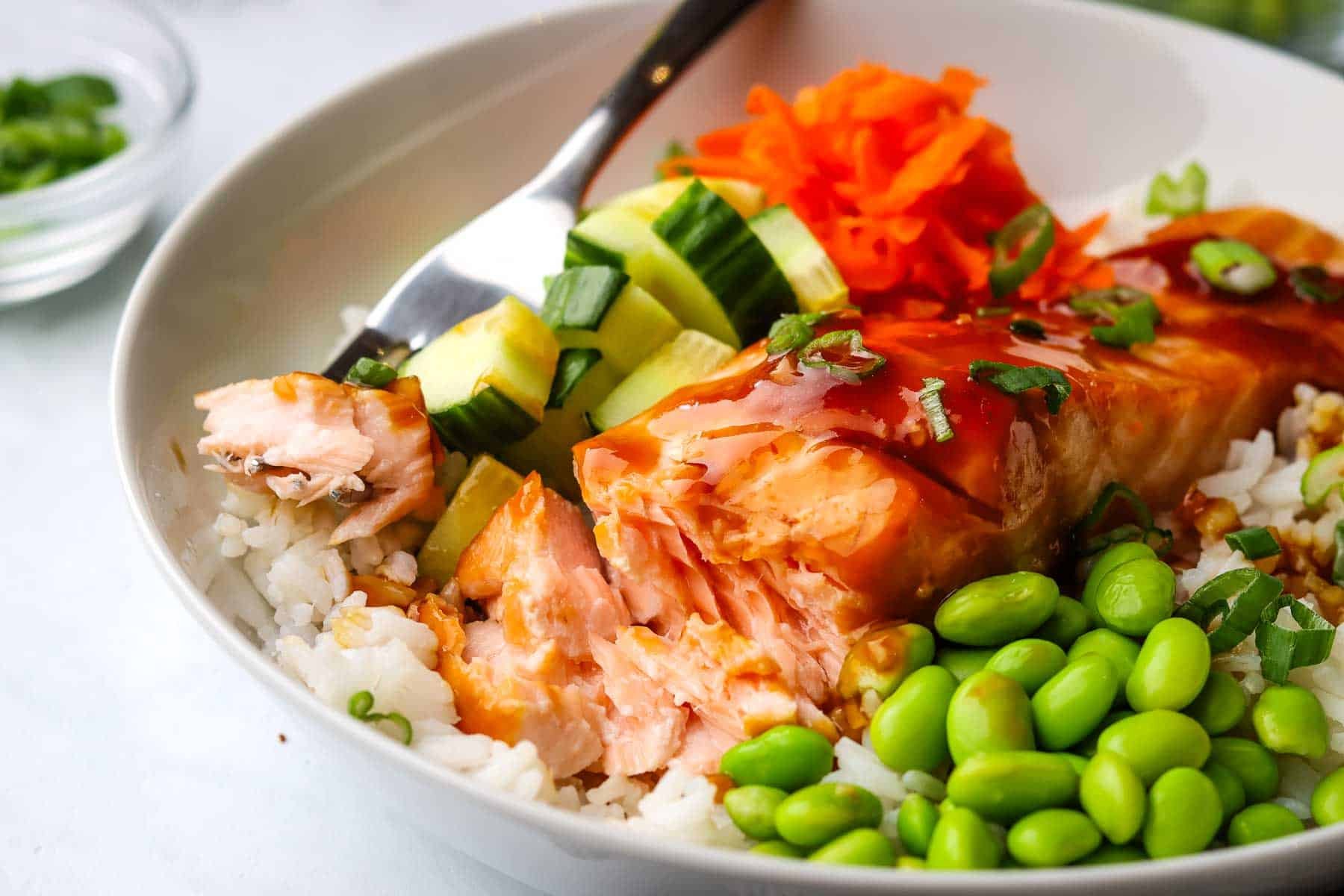 a fork taking a bite of the salmon from a bowl with rice, salmon, veggies, and sauce.
