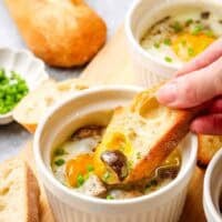 A ramekin with a baked egg in it with a hand dipping a baguette slice into the yolk.