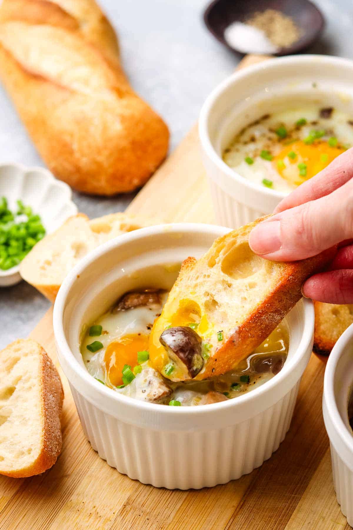 A ramekin with a baked egg in it with a hand dipping a baguette slice into the yolk.
