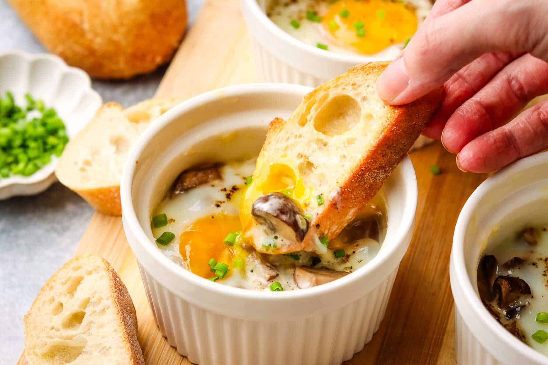 A ramekin with a baked egg in it with a hand dipping a baguette slice into the yolk.