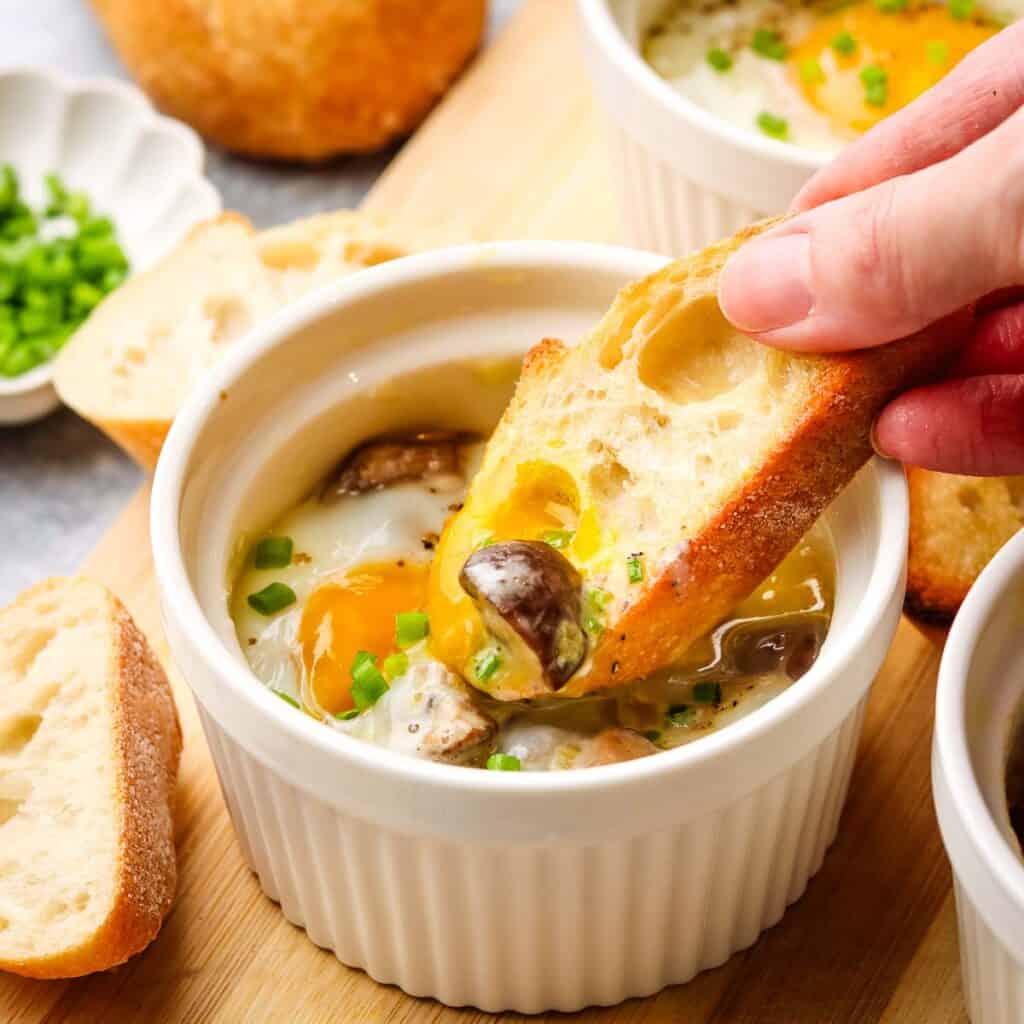 A ramekin with a baked egg in it with a hand dipping a baguette slice into the yolk.