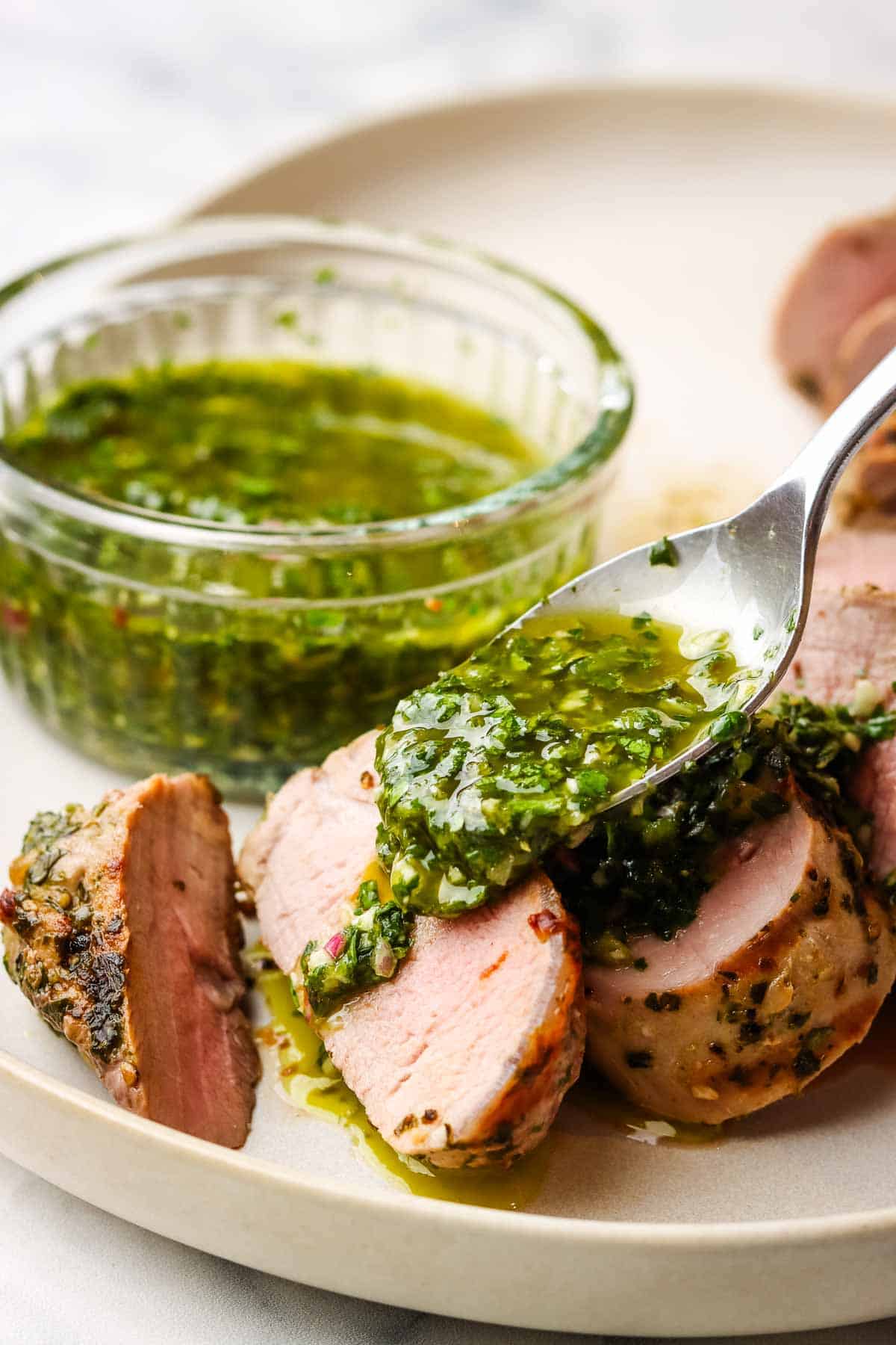 Spooning chimichurri over a plate of sliced pork tenderloin.