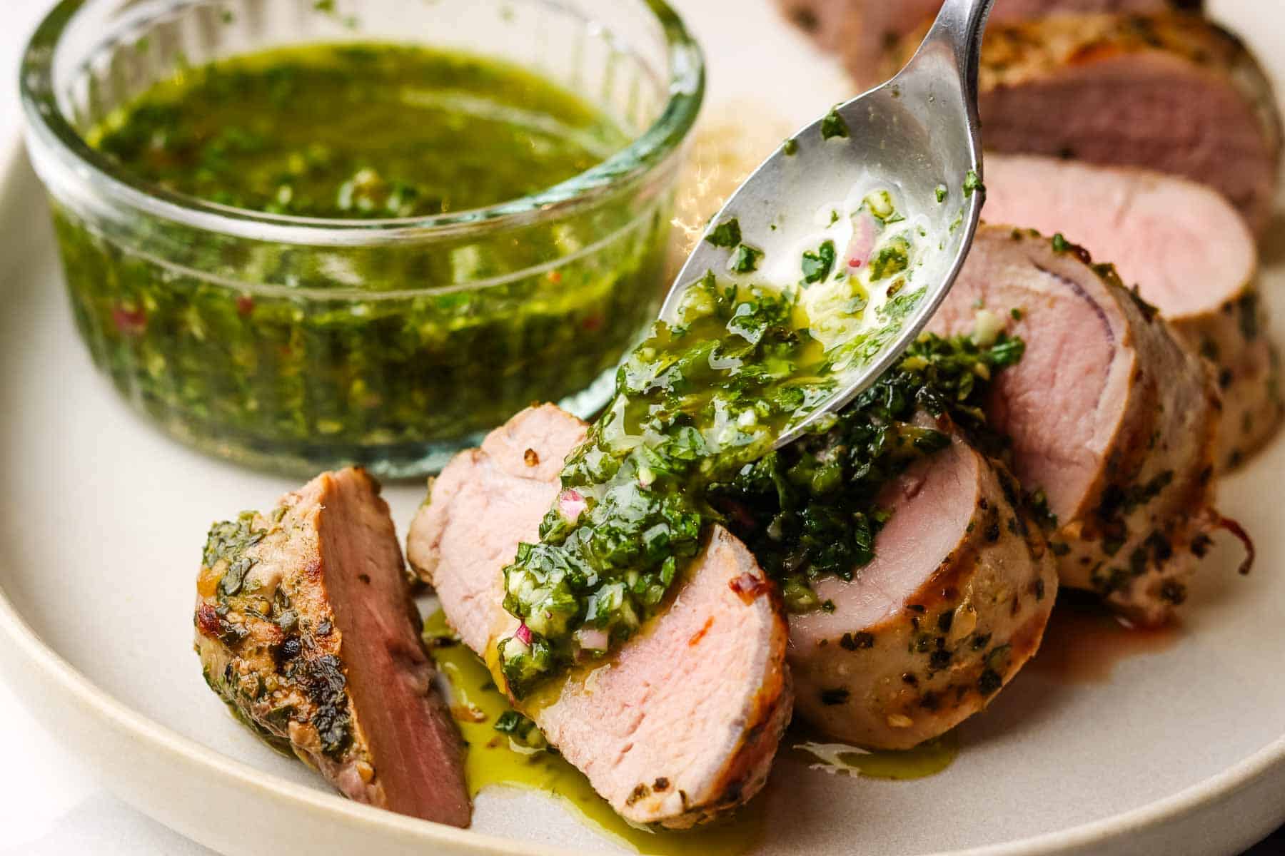 Spooning chimichurri over a plate of sliced pork tenderloin.