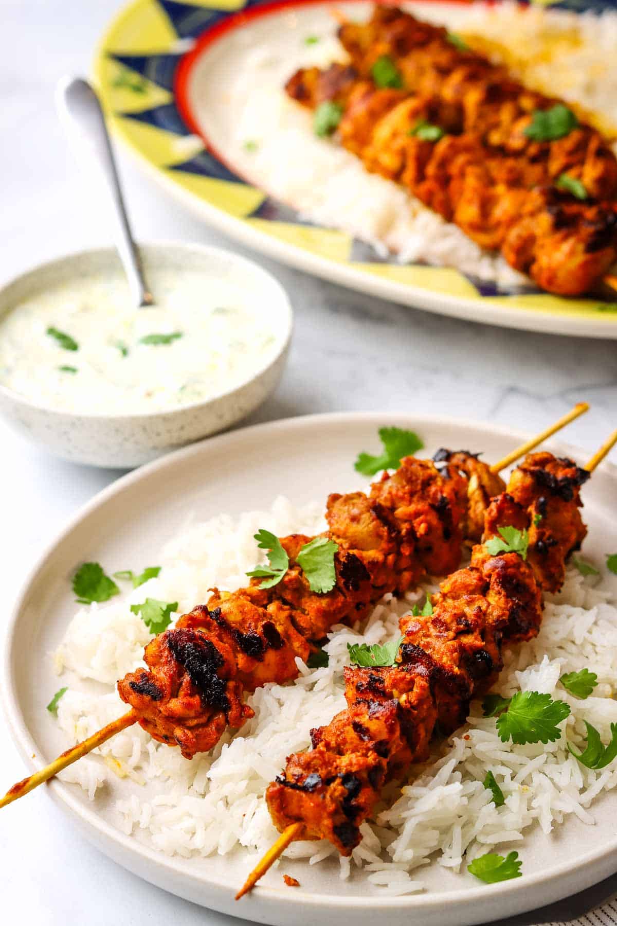 a plate of tandoori chicken skewers served on a bed of white rice with raita dipping sauce in a small bowl.