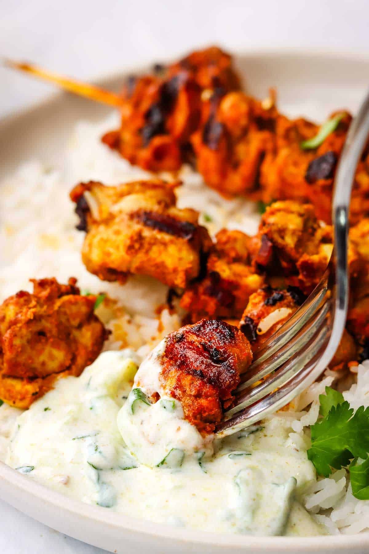 picking up a bite of tandoori chicken skewers dipped in raita and with white rice from a plate.