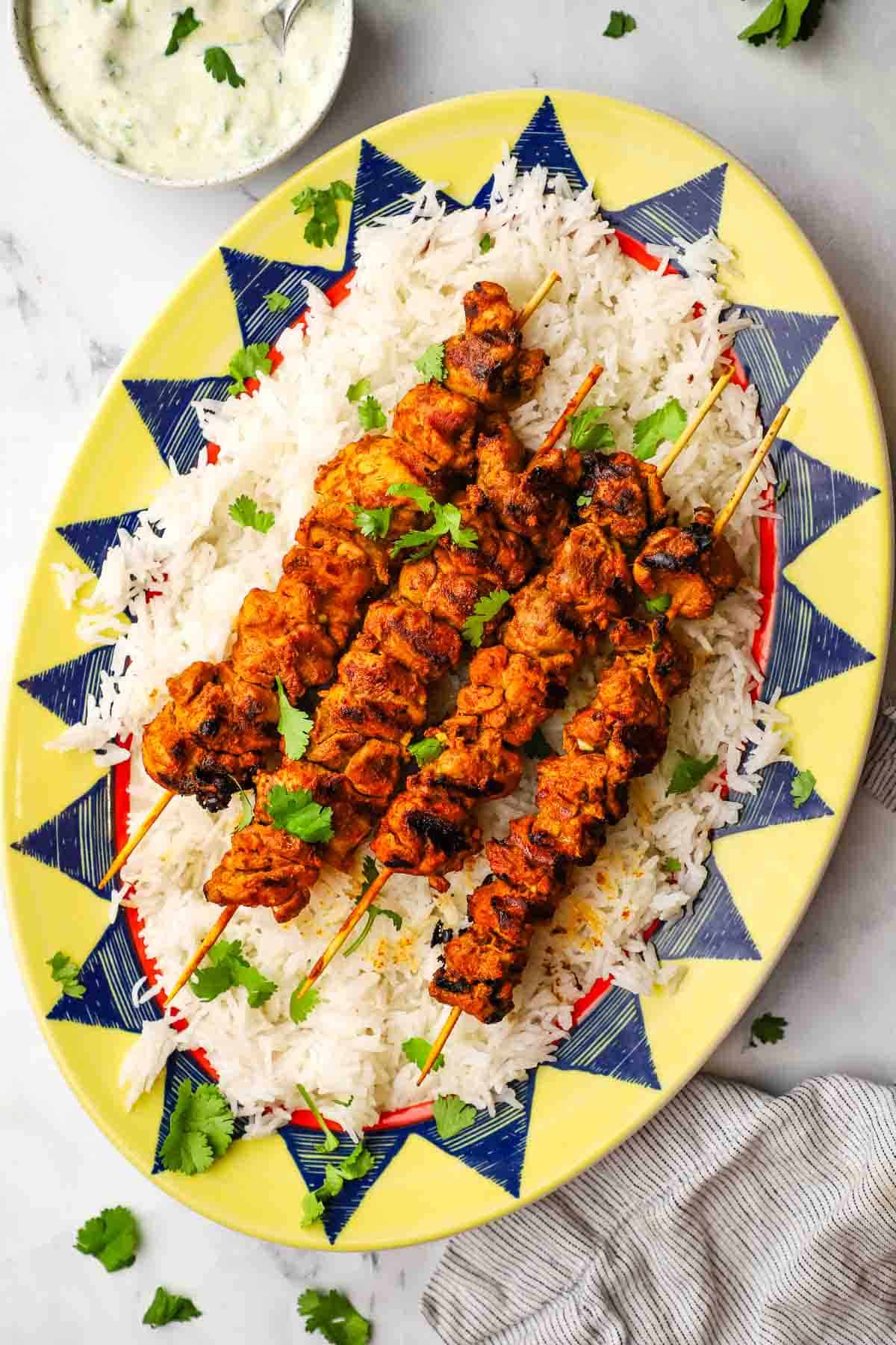 a platter of white rice with tandoori chicken skewers on top and raita on the side