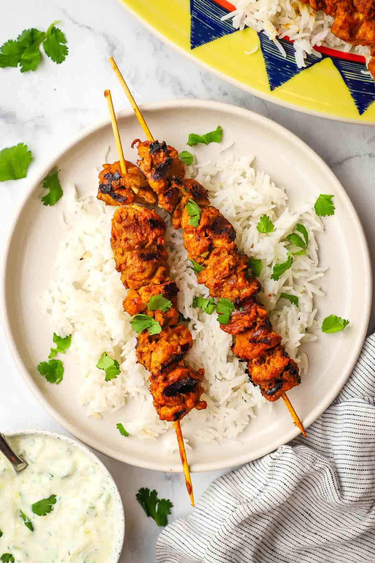 a plate of tandoori chicken skewers served on a bed of white rice with raita dipping sauce in a small bowl.