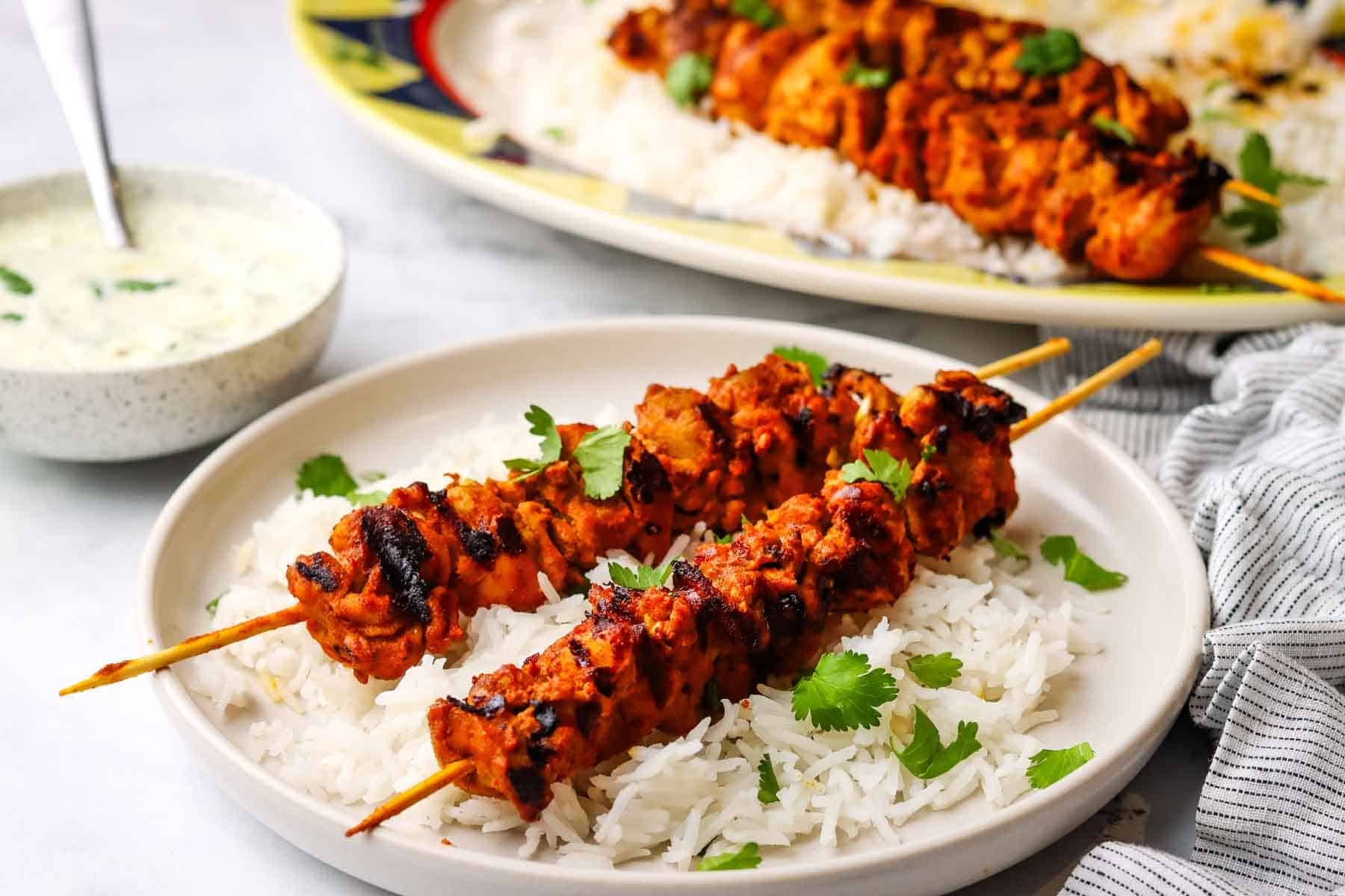 a plate of tandoori chicken skewers served on a bed of white rice with raita dipping sauce in a small bowl.