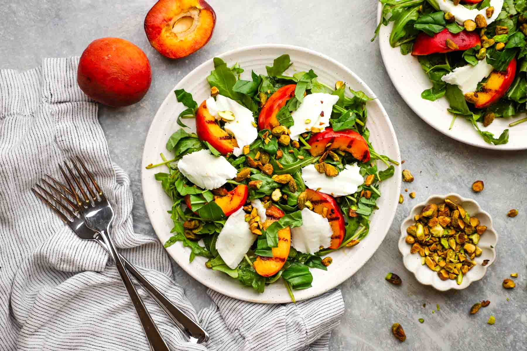 Grilled peach salad with arugula, fresh mozzarella, toasted pistachios, and herbs, lightly dressed and served on a plate.