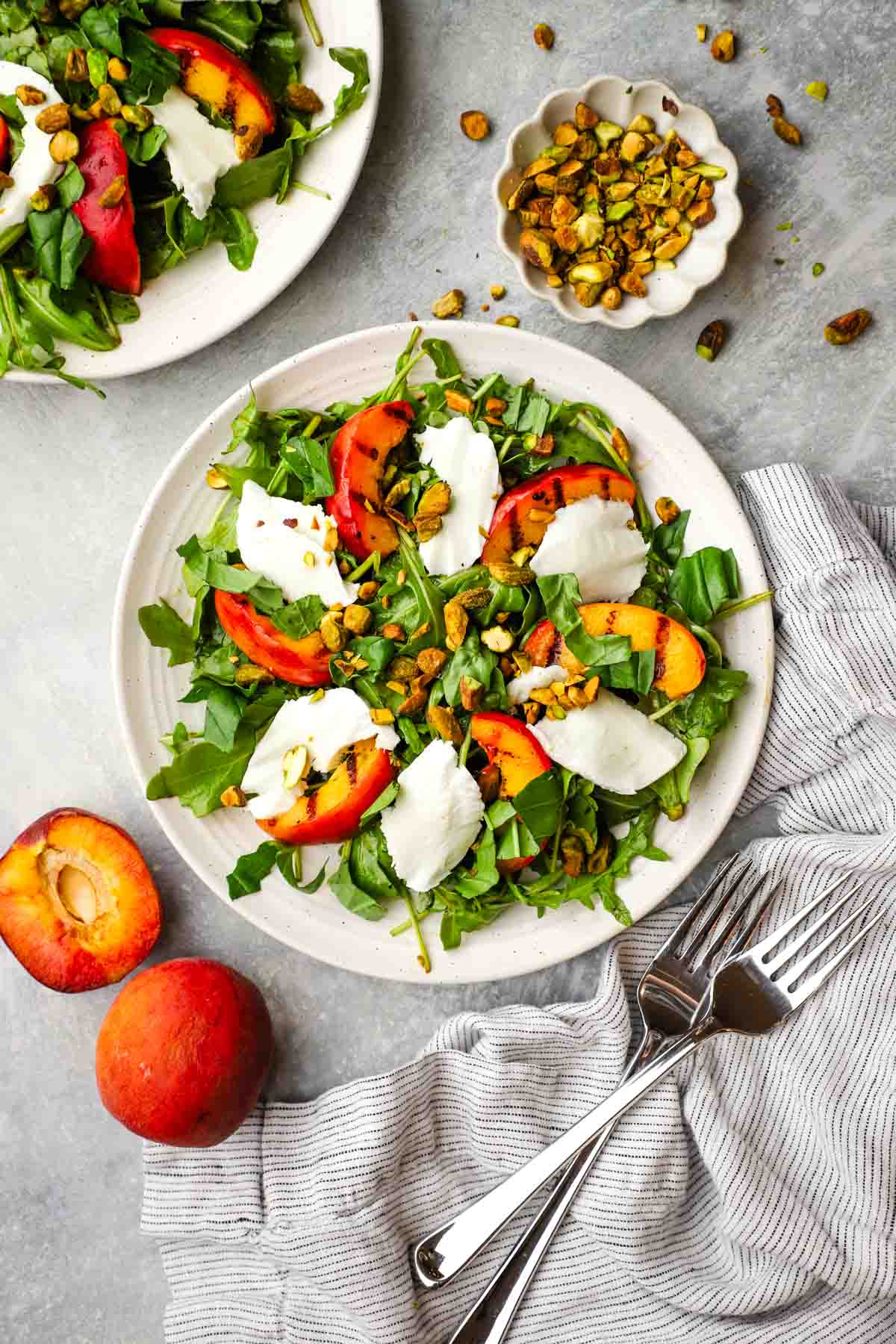 Grilled peach salad with arugula, fresh mozzarella, toasted pistachios, and herbs, lightly dressed and served on a plate.