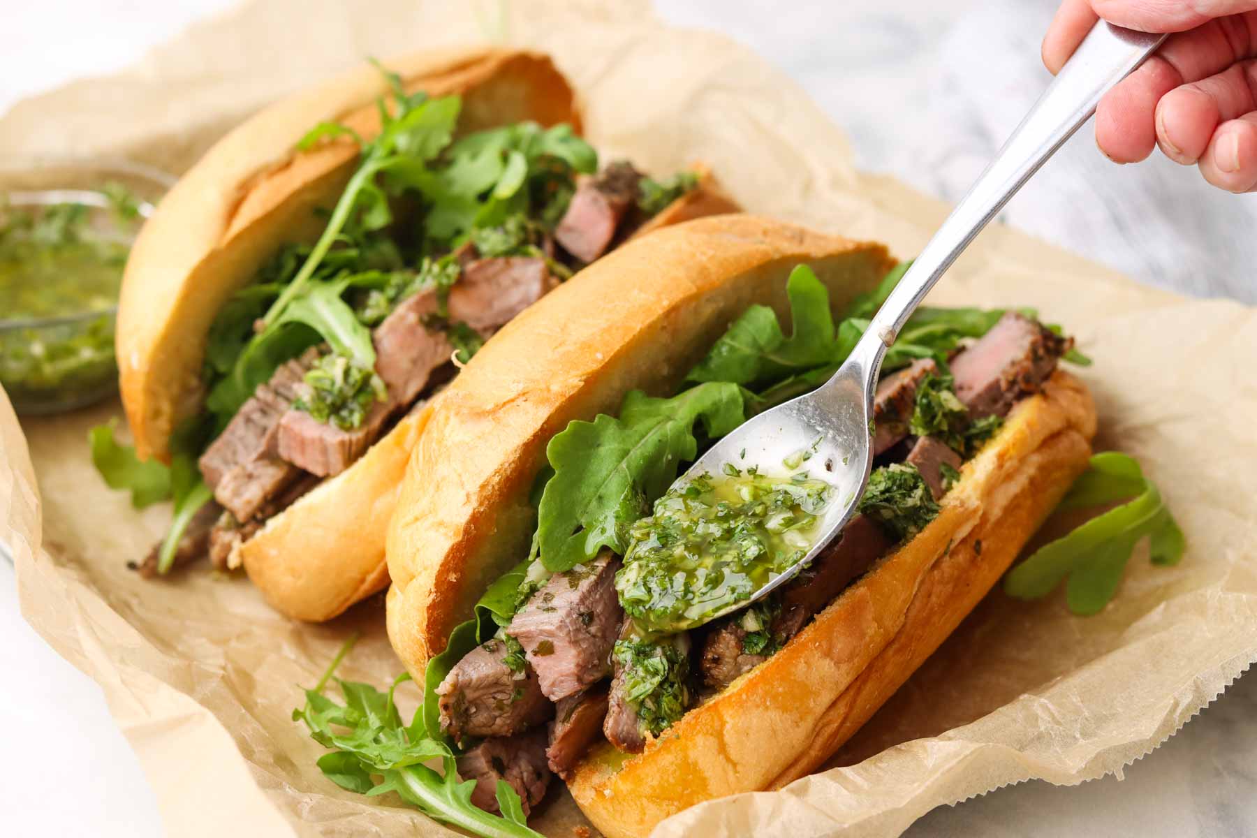 adding chimichurri with a spoon on a sandwich on a platter