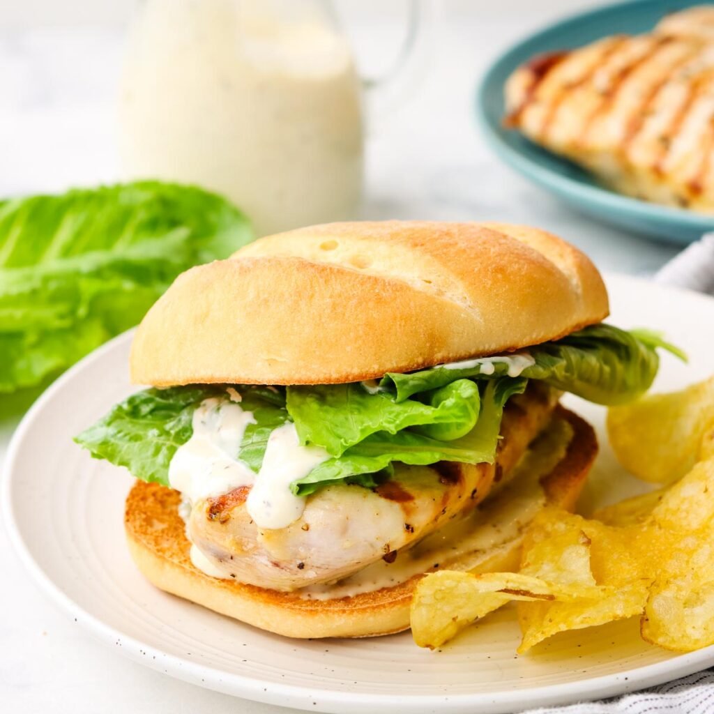 A chicken Caesar sandwich on a plate with potato chips on the side.