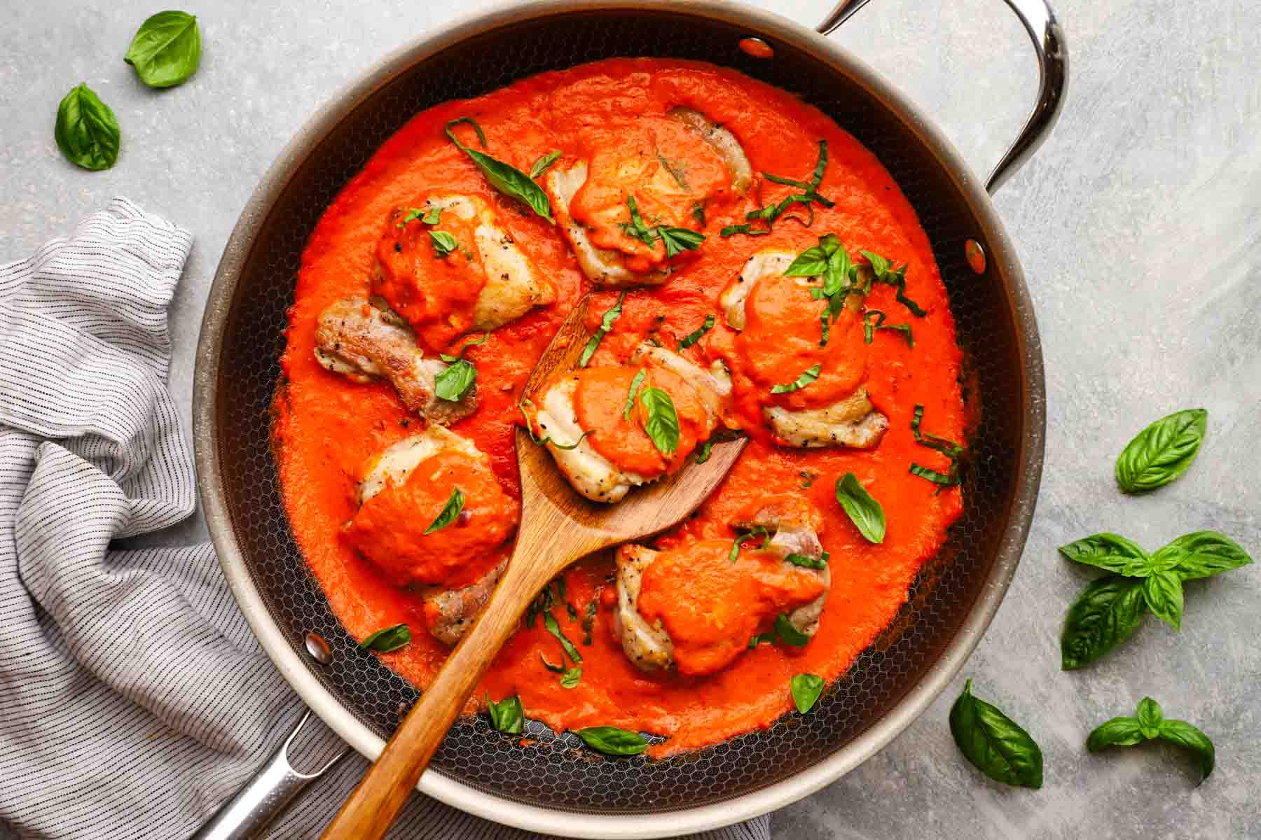 a skillet with chicken thighs in a roasted red pepper sauce and a wooden spoon in it for serving