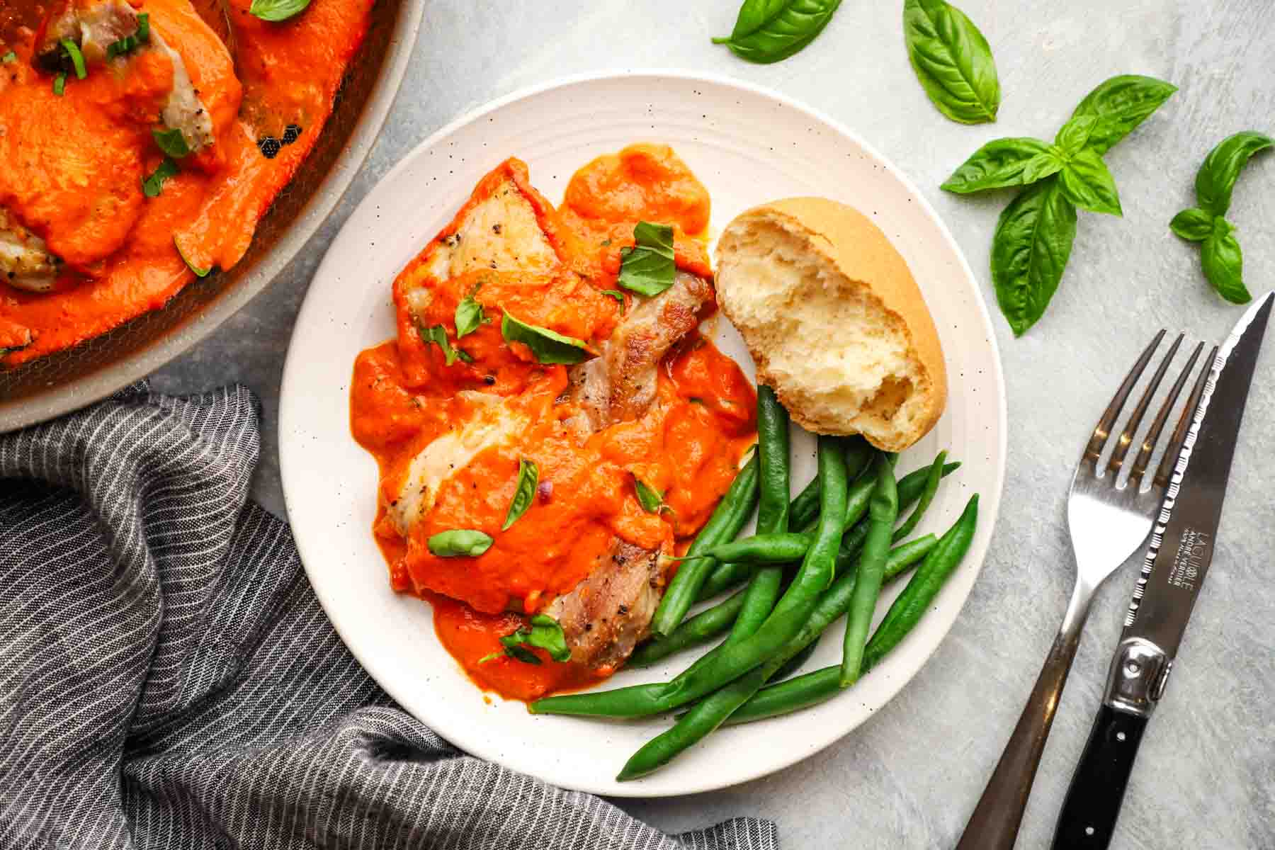 overhead view of a plate of roasted red pepper chicken served with green beans and a slice of bread.