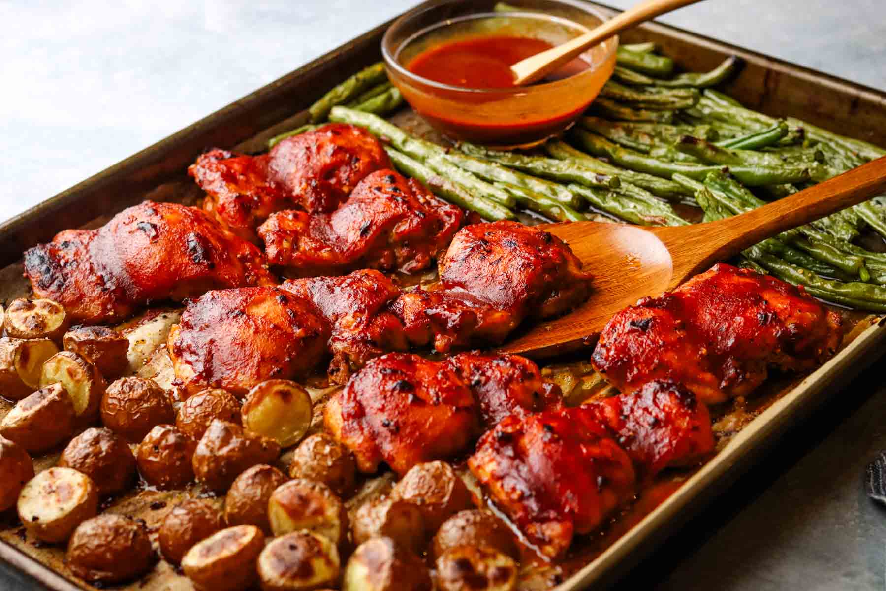 a baking dish with chicken thighs, green beans, and roasted potatoes with some sauce in a bowl.