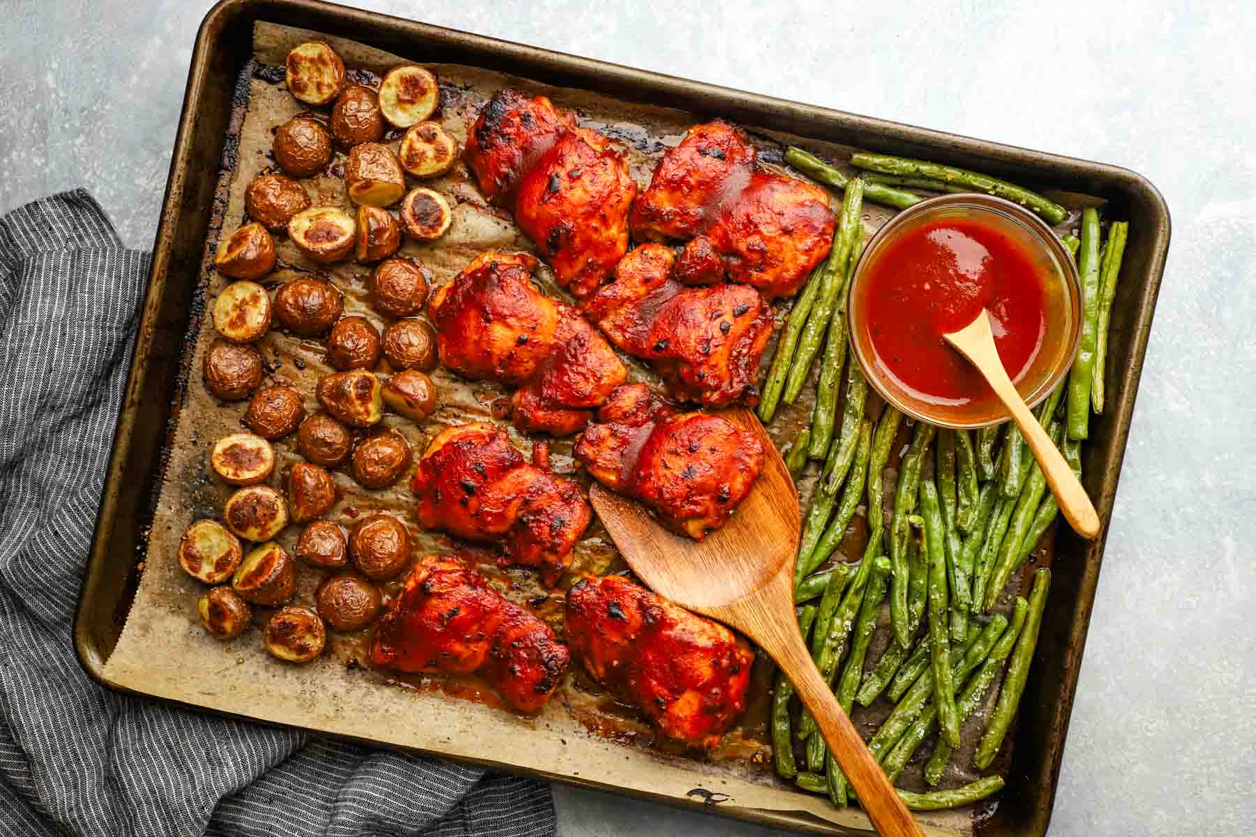 overhead view of a baking sheet with chicken, potatoes, green beans, and a bowl of sauce.