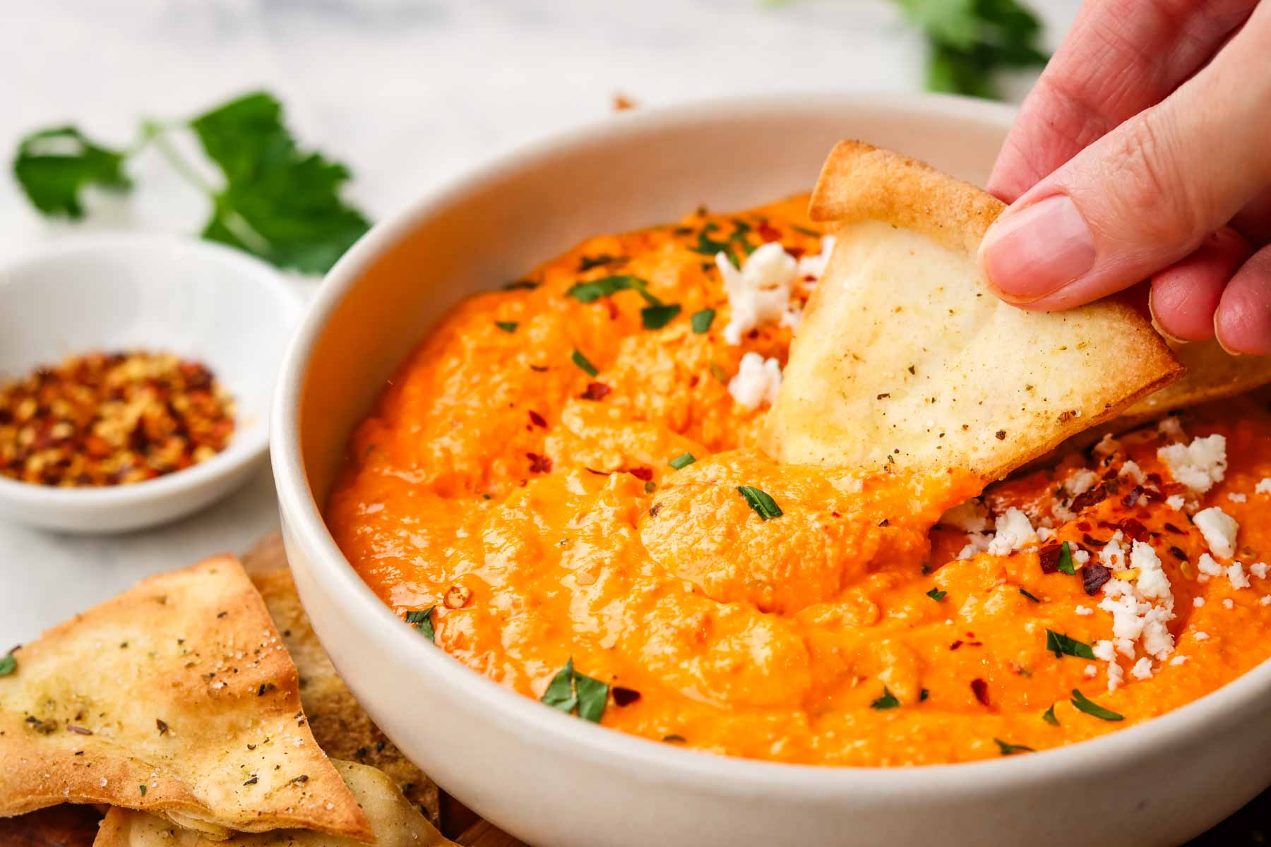 a bowl of feta and roasted red pepper dip with crunchy pita dipped in the bowl and more on the side