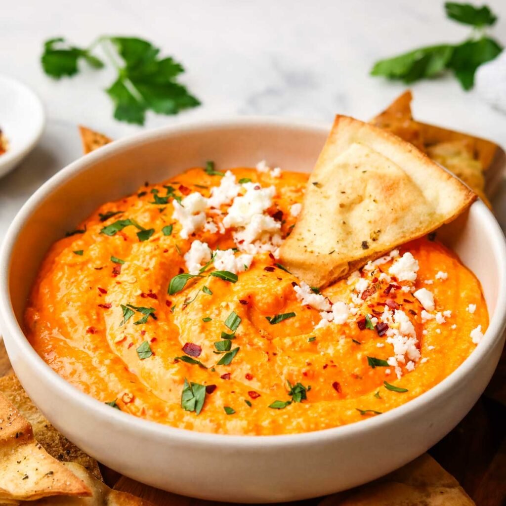 a bowl of feta and roasted red pepper dip with a crunchy pita on the side