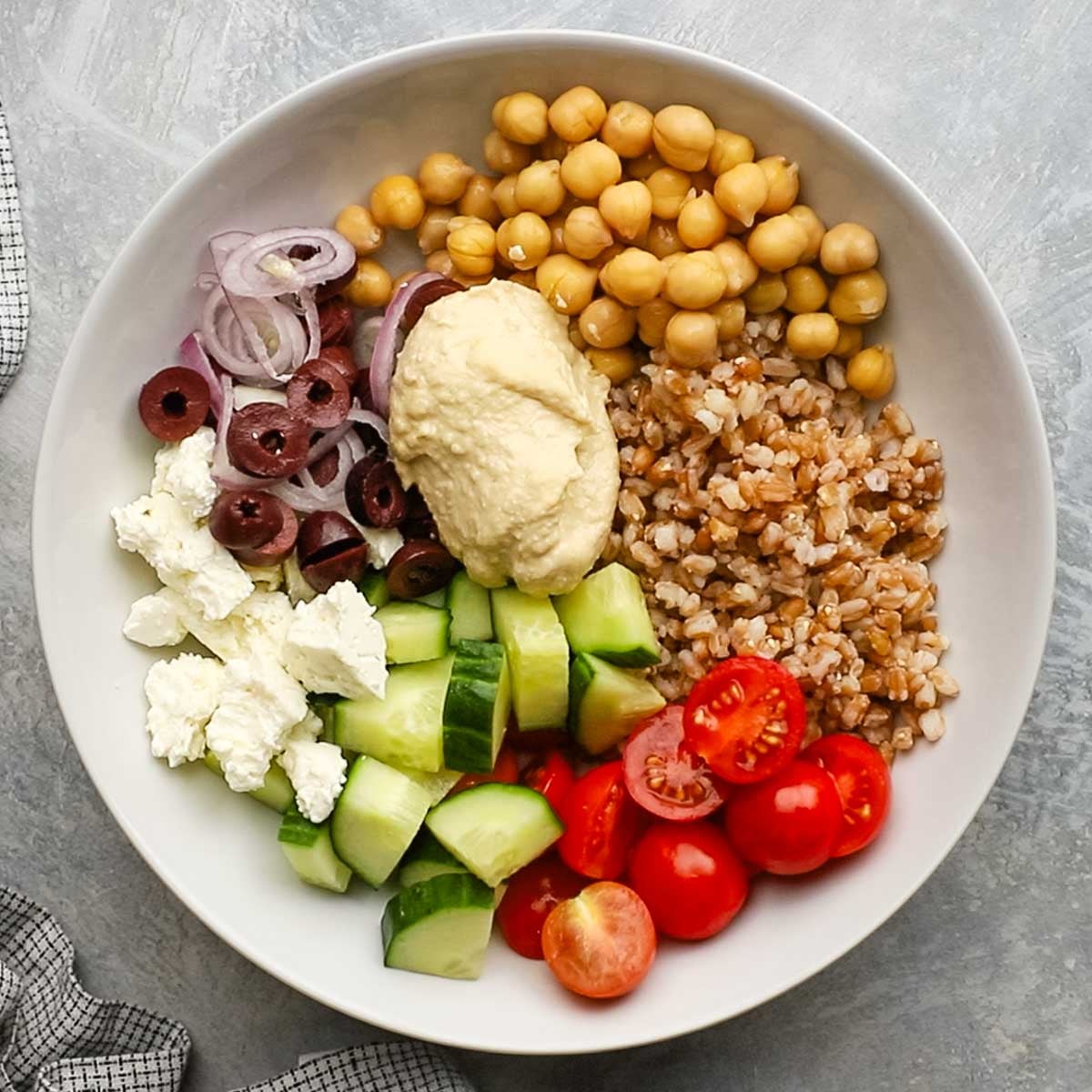 a bowl with farro, chickpeas,fresh chopped vegetables, feta cheese, olives, and hummus