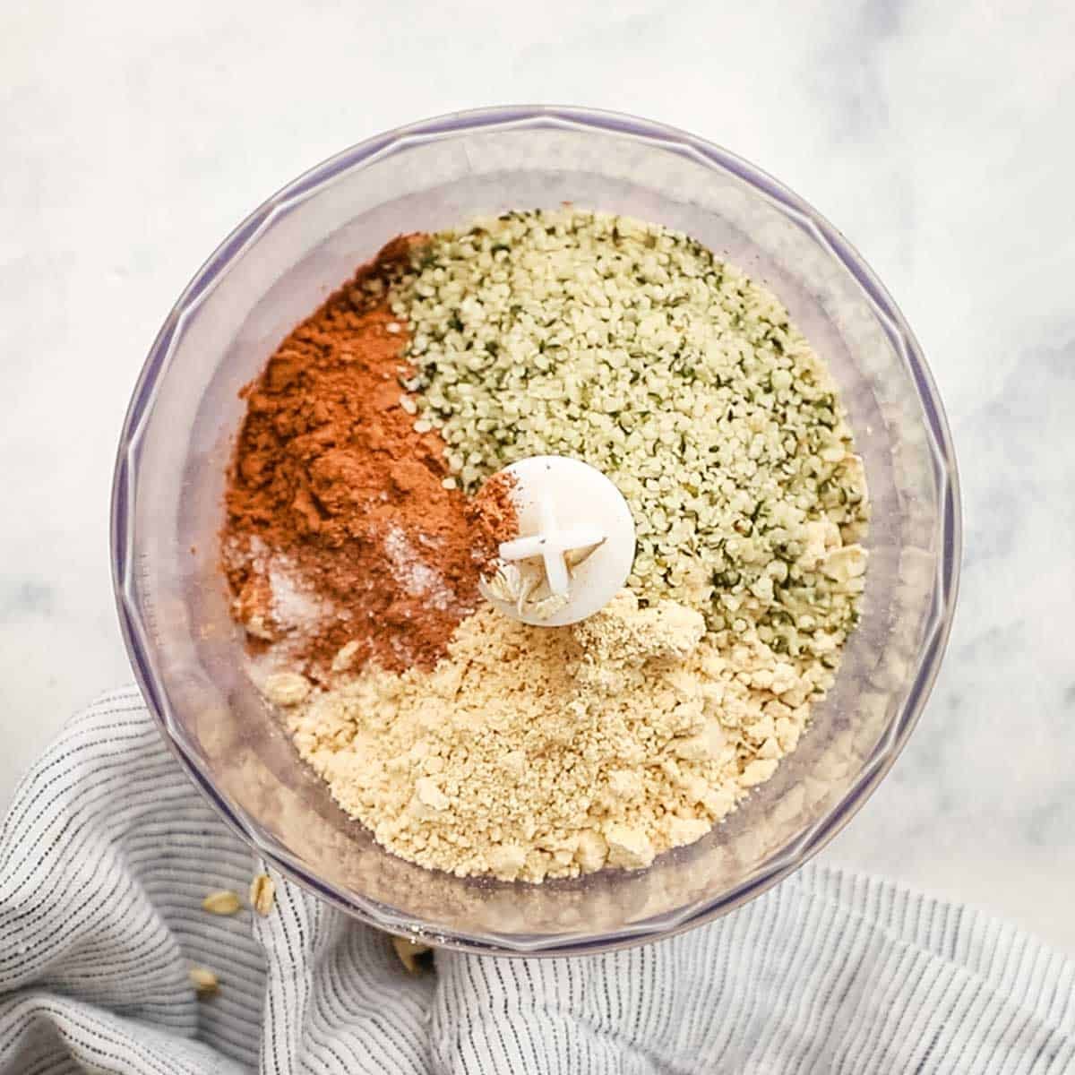 Rolled oats, peanut butter powder, cocoa powder, hemp seeds, and more in a food processor ready to be blended. 