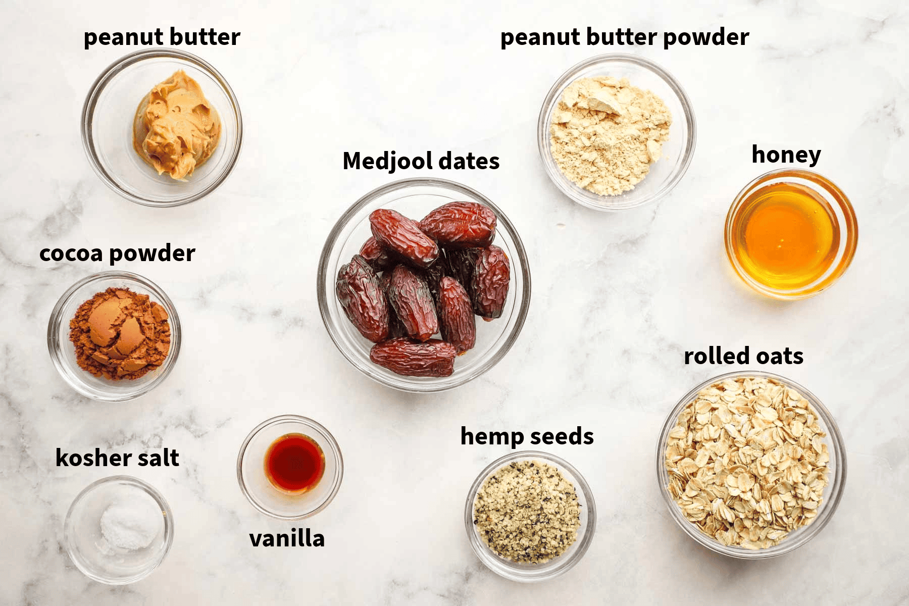 Top-down view of protein ball ingredients placed in individual bowls that include medjool dates, peanut butter, peanut butter powder, hemp seeds, vanilla, rolled oats, and more. 