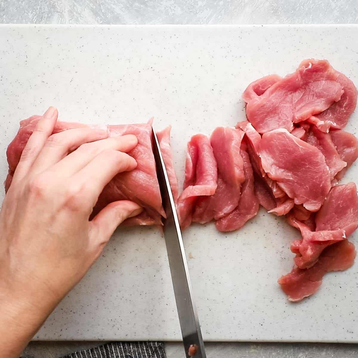 Top-down view of raw pork tenderloin being cut into thin slices with a sharp knife on a cutting board for stir-fry.