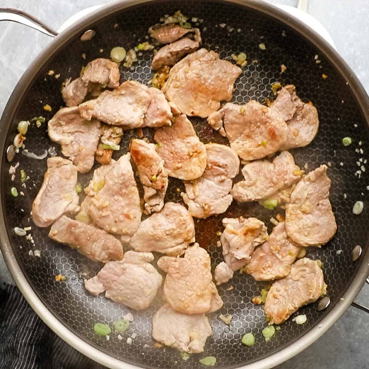Top-down view of pork tenderloin being cooked in a pan with a small amount of oil, scallions, and garlic.
