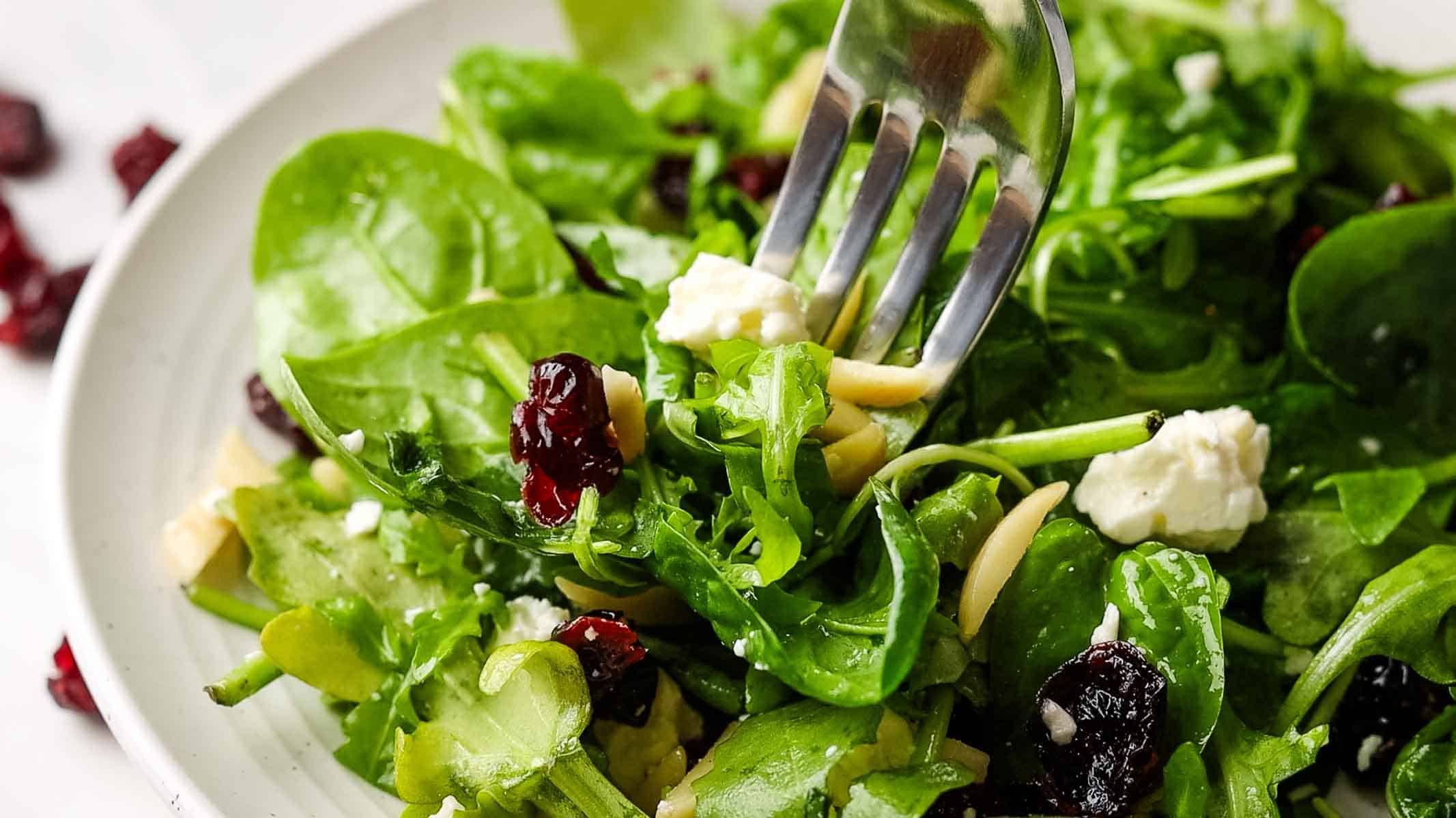 Close up of spinach arugula with sliced almonds, dried cranberries, and feta with a fork, readying a bite from the salad bowl.